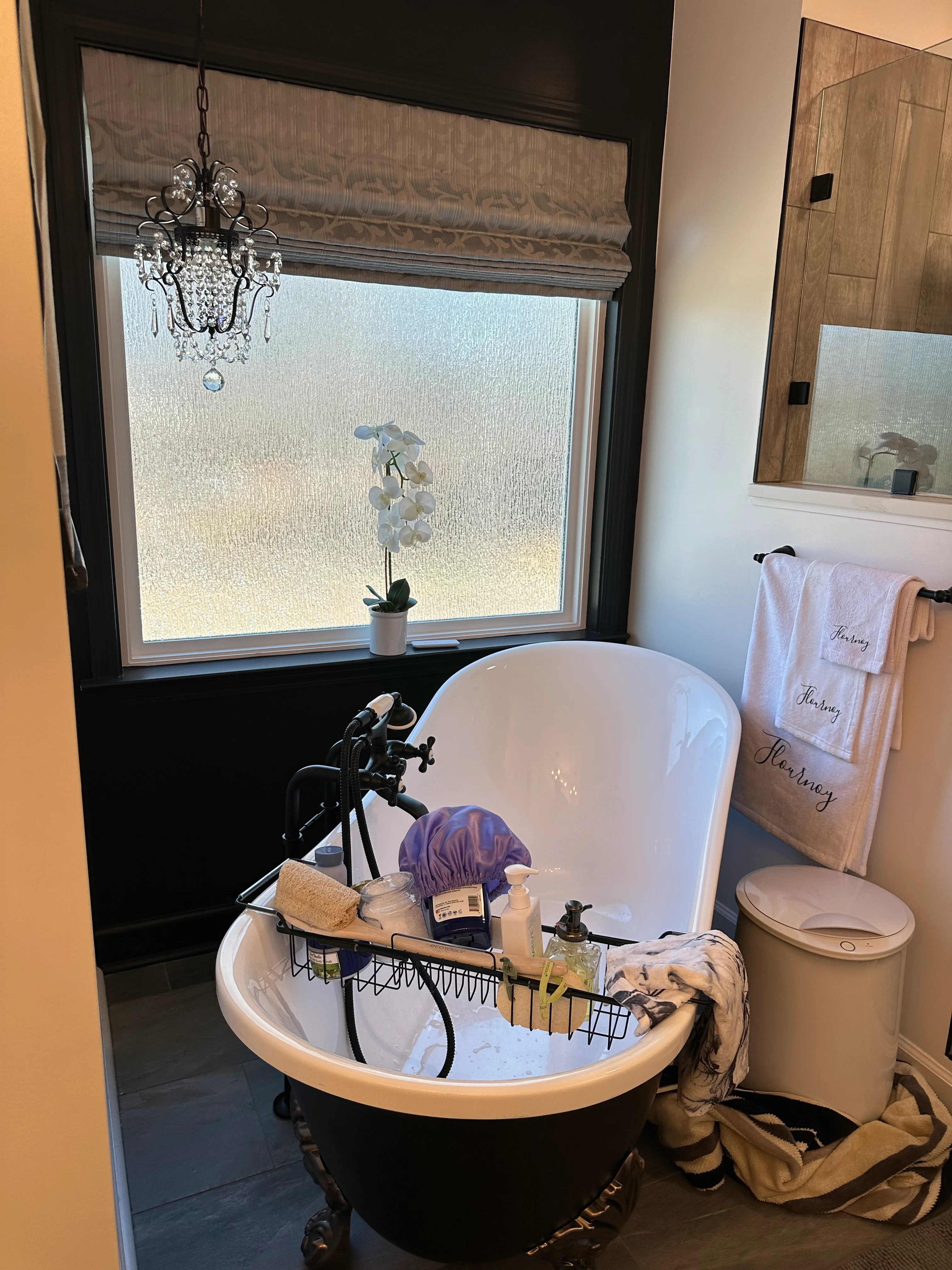 Modern bathroom with black and white freestanding tub, crystal chandelier, and window with Roman shade